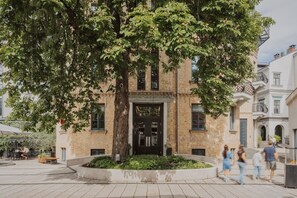 A building with a large tree in front, a sign reading 'Viesca Hotel Bergs,' and a paved plaza with people walking. - Hotel Bergs Suites (Riga)