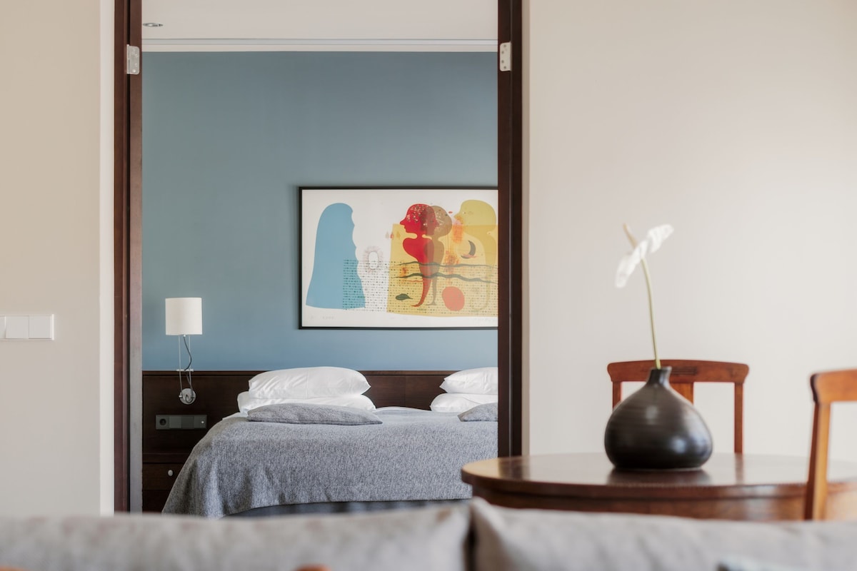 A hotel room with a bed, a bedside table with a vase, a wall-mounted lamp, and a colorful abstract painting.