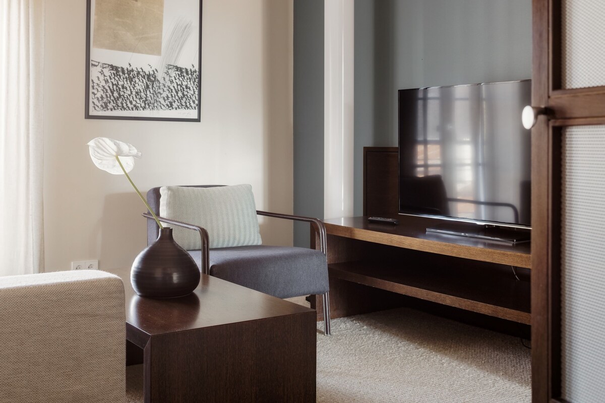 A modern living room with a flat-screen TV, a dark wooden coffee table, a vase with a plant, and a grey armchair.