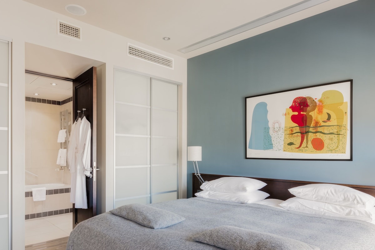 A hotel room with a bed, a nightstand, a wall-mounted lamp, a framed abstract painting, and a walk-in closet with hanging robes.