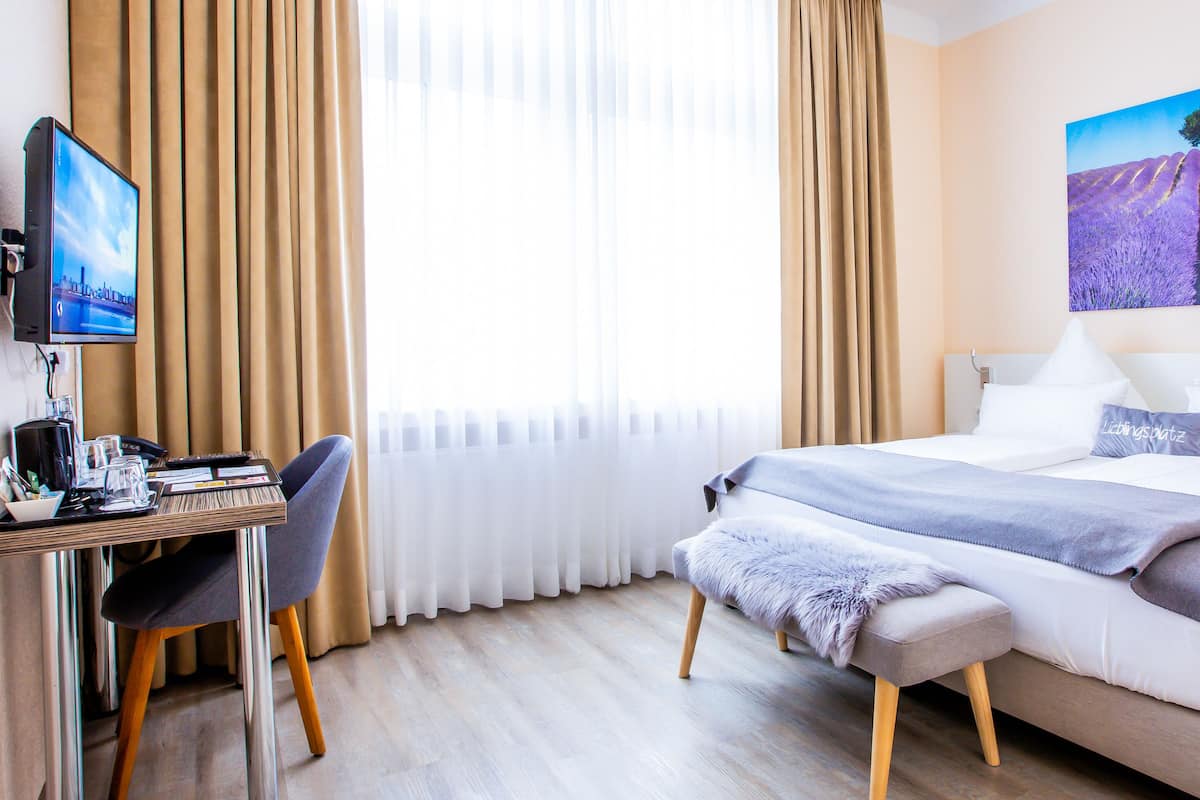 A hotel room with a bed, a desk with a computer, a chair, a window with curtains, and a painting on the wall.
