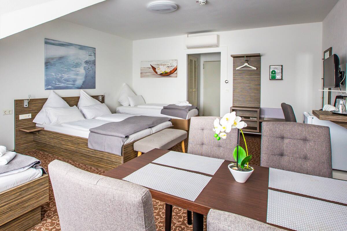 A hotel room with two beds, a dining table with chairs, a small table with a potted plant, and a TV on a stand.