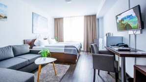 Une chambre d’hôtel moderne équipée d’un canapé, d’une petite table avec une plante, d’un lit donnant sur l’extérieur, d’un bureau avec un ordinateur et d’un téléviseur fixé au mur.