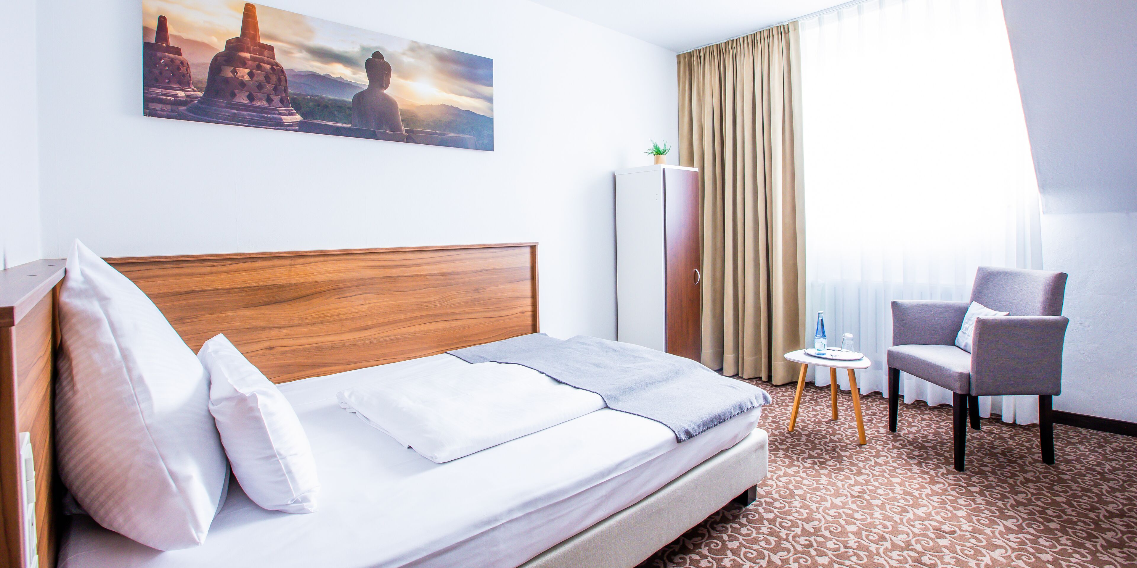 Een hotelkamer met een bed, een stoel, een tafeltje, een kledingkast en een schilderij aan de muur.