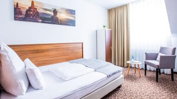 A hotel room with a bed, a chair, a small table, a wardrobe, and a painting on the wall.