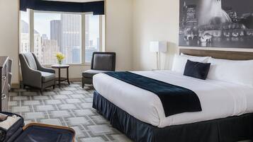 A hotel room with a large bed, a desk, two chairs, a small table with flowers, a suitcase, and a cityscape view.