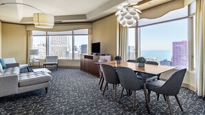 A hotel room with a large window, a dining area with a table and chairs, a sofa, a TV, and a decorative light fixture. - The Buckingham Hotel (Chicago)