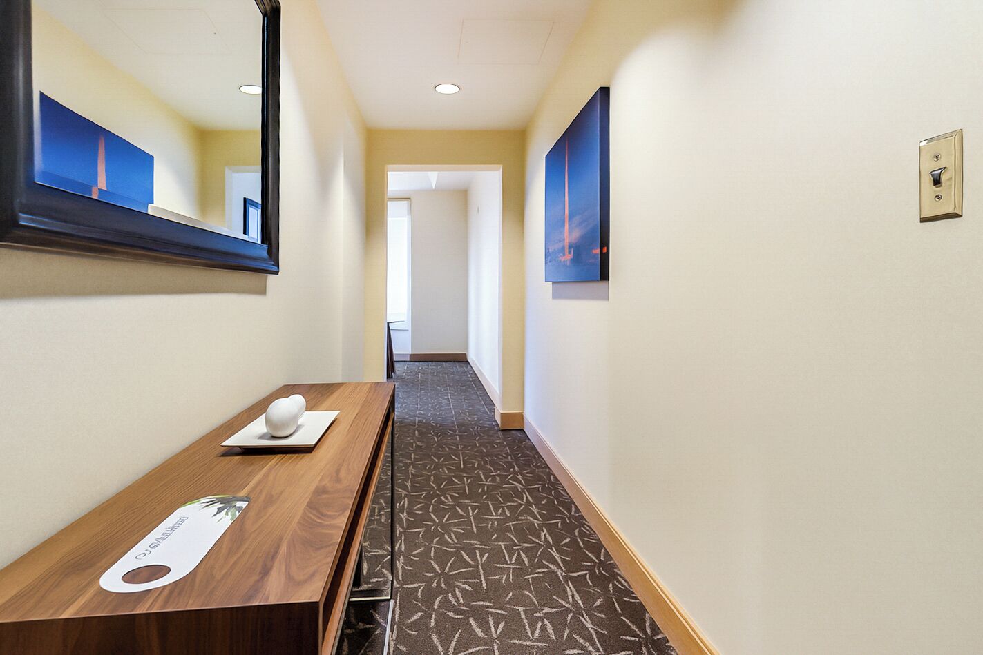 Een hotelgang met een houten receptiebalie, een spiegel en twee kunstwerken in lijsten aan de muur.