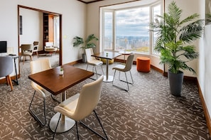 A modern hotel room with a city view, a dining area with a table and chairs, and a large potted plant.