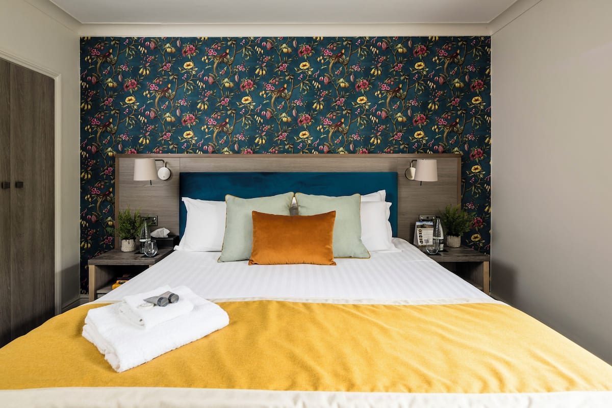 Cama bem arrumada com manta amarela, cabeceira azul e papel de parede floral.