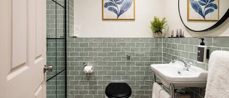 Un baño con inodoro, lavamanos y ducha. Hay dos cuadros con hojas enmarcadas en la pared.