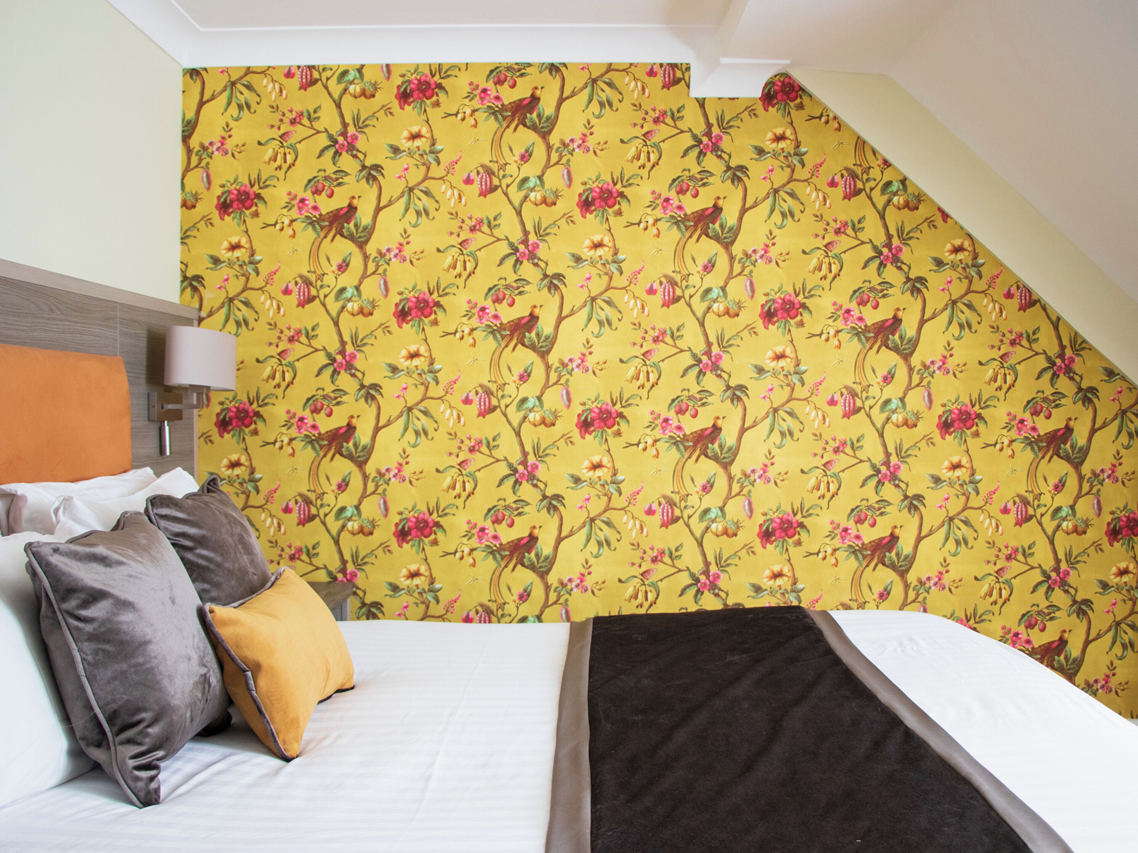 Un dormitorio con cama, almohadas, una lámpara y papel tapiz con motivos florales.