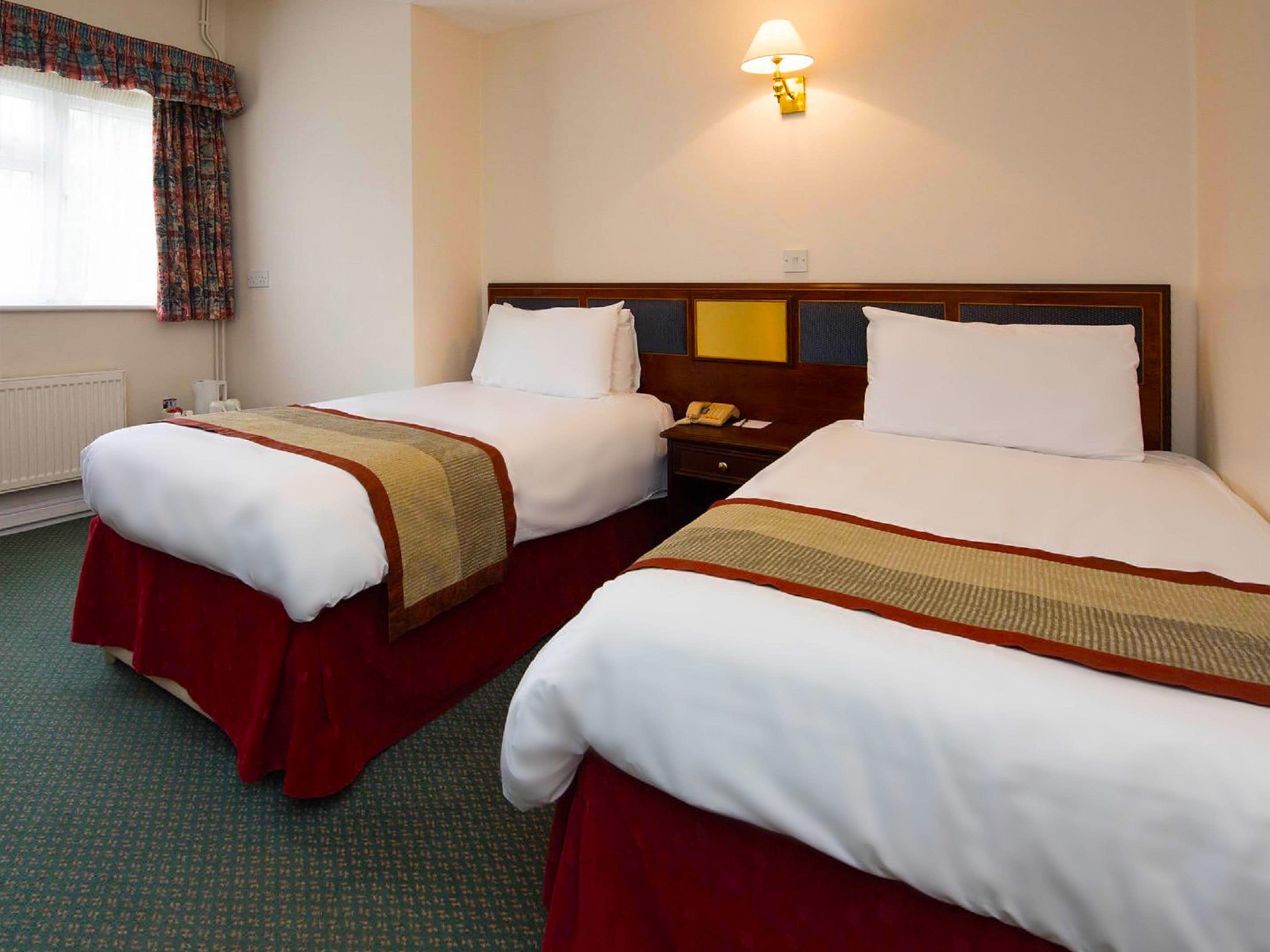 A hotel room with two beds, a bedside table with a lamp, and a window with curtains.