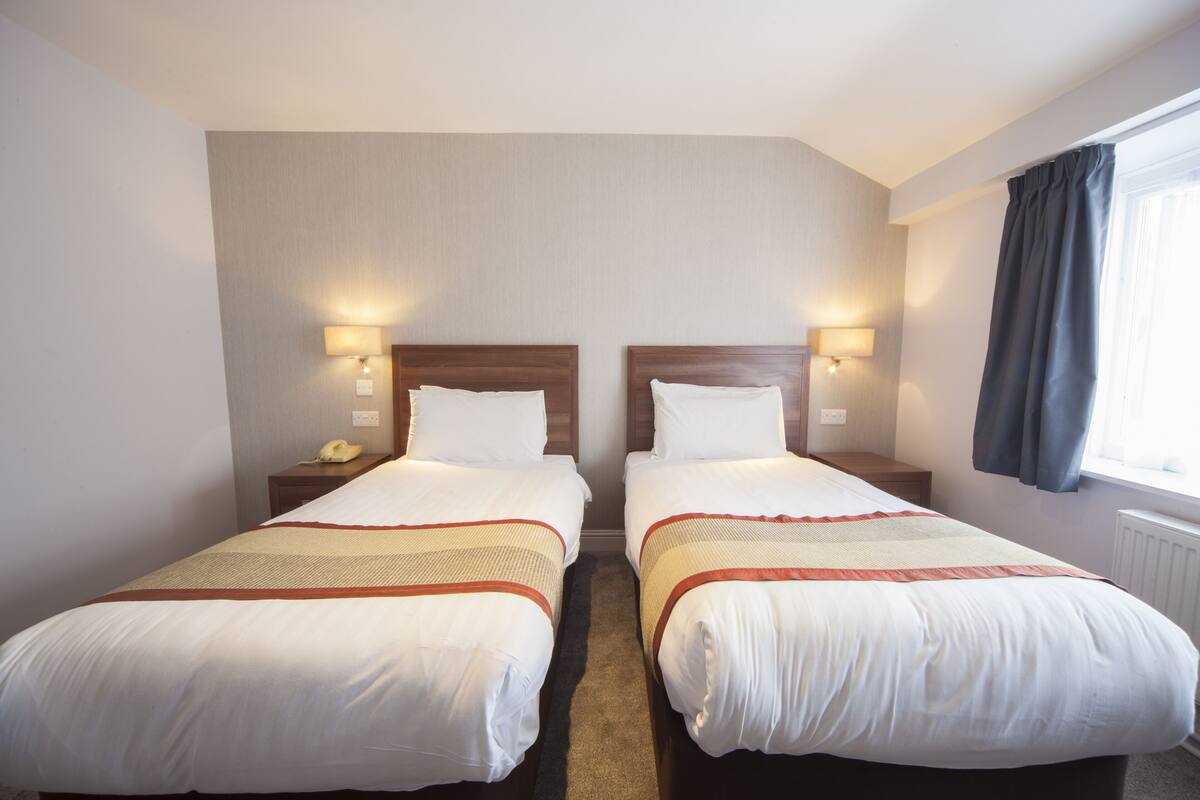 A hotel room with two beds, each with a wooden headboard, white bedding, and a red and beige striped blanket. There are bedside lamps, a telephone, and a window with curtains.