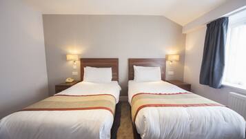A hotel room with two beds, each with a wooden headboard, white bedding, and a red and beige striped blanket. There are bedside lamps, a telephone, and a window with curtains.