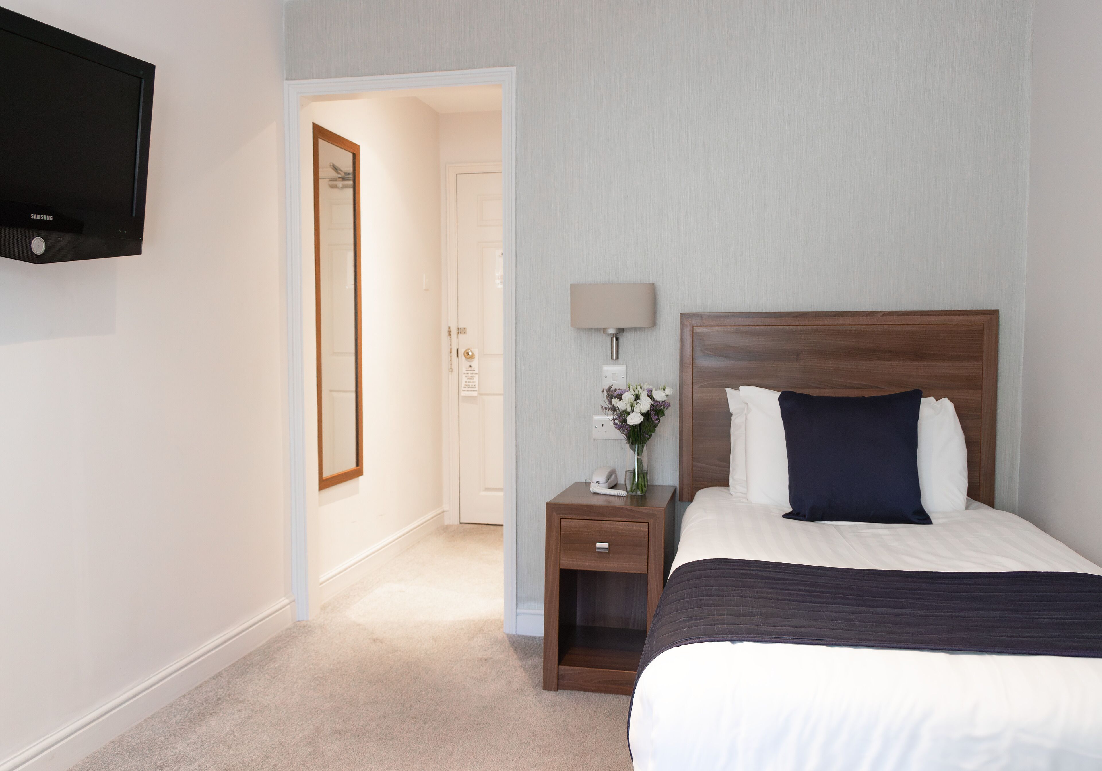 A hotel room with a bed, a nightstand, a lamp, and a TV mounted on the wall.