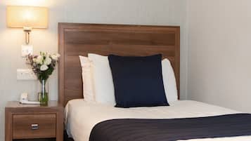 Una cama bien hecha con almohadas blancas y azul marino, un cabecero oscuro y una mesita de noche con una lámpara y un florero.