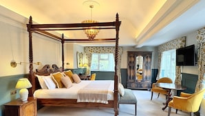 A four-poster bed with a chandelier, a small table with a lamp, a chair, and a television on the wall. - The Holt Hotel (Bicester)
