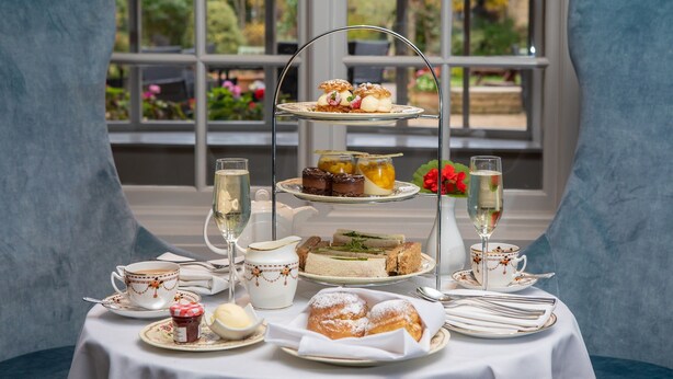 A three-tiered dessert stand with various pastries and cakes, a pitcher of cream, a teapot, and a cup of tea on a table with a white tablecloth. - The Holt Hotel (Bicester)