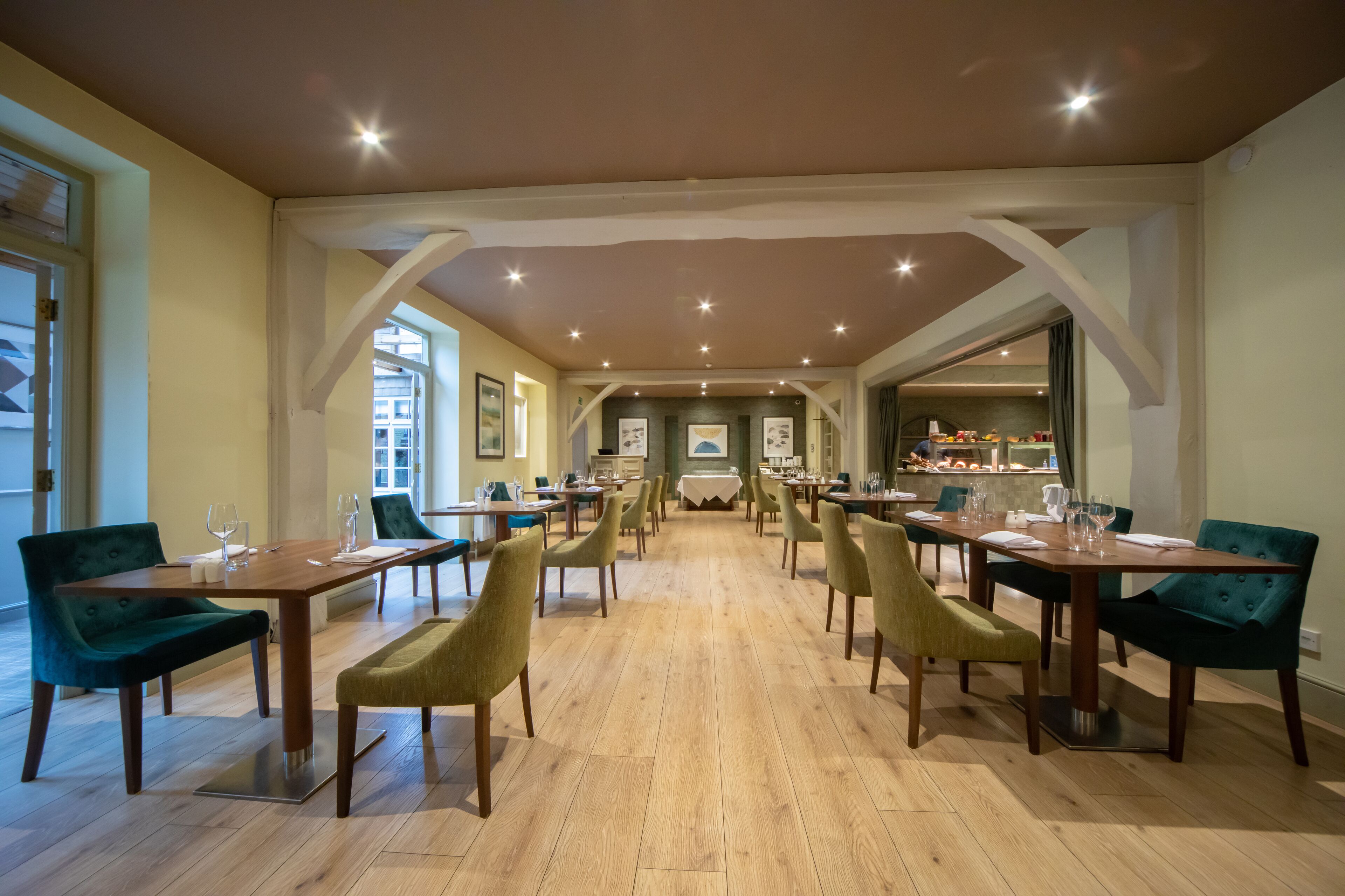 A spacious dining area with multiple tables and chairs, wooden flooring, and a view of the kitchen in the background.