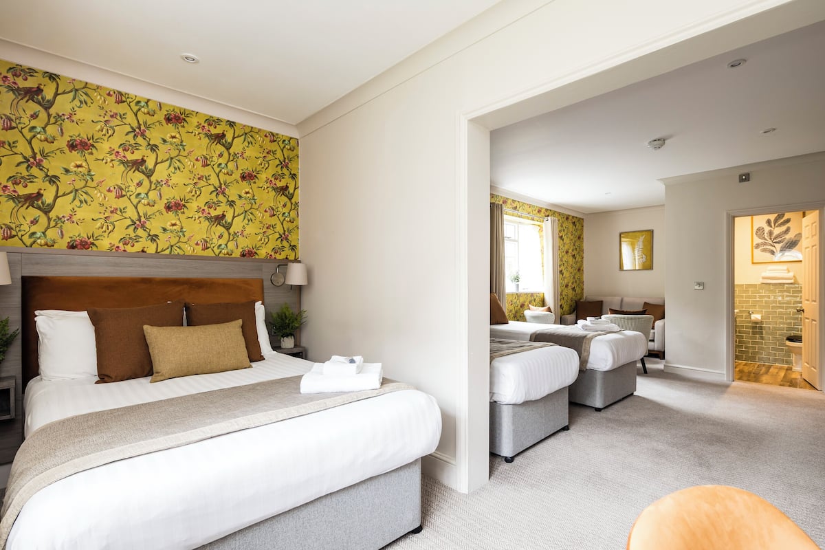 A hotel room with a large bed, a smaller bed, and a sofa. There is a bathroom with a shower and a toilet. The room has a yellow floral wallpaper and a wooden headboard.