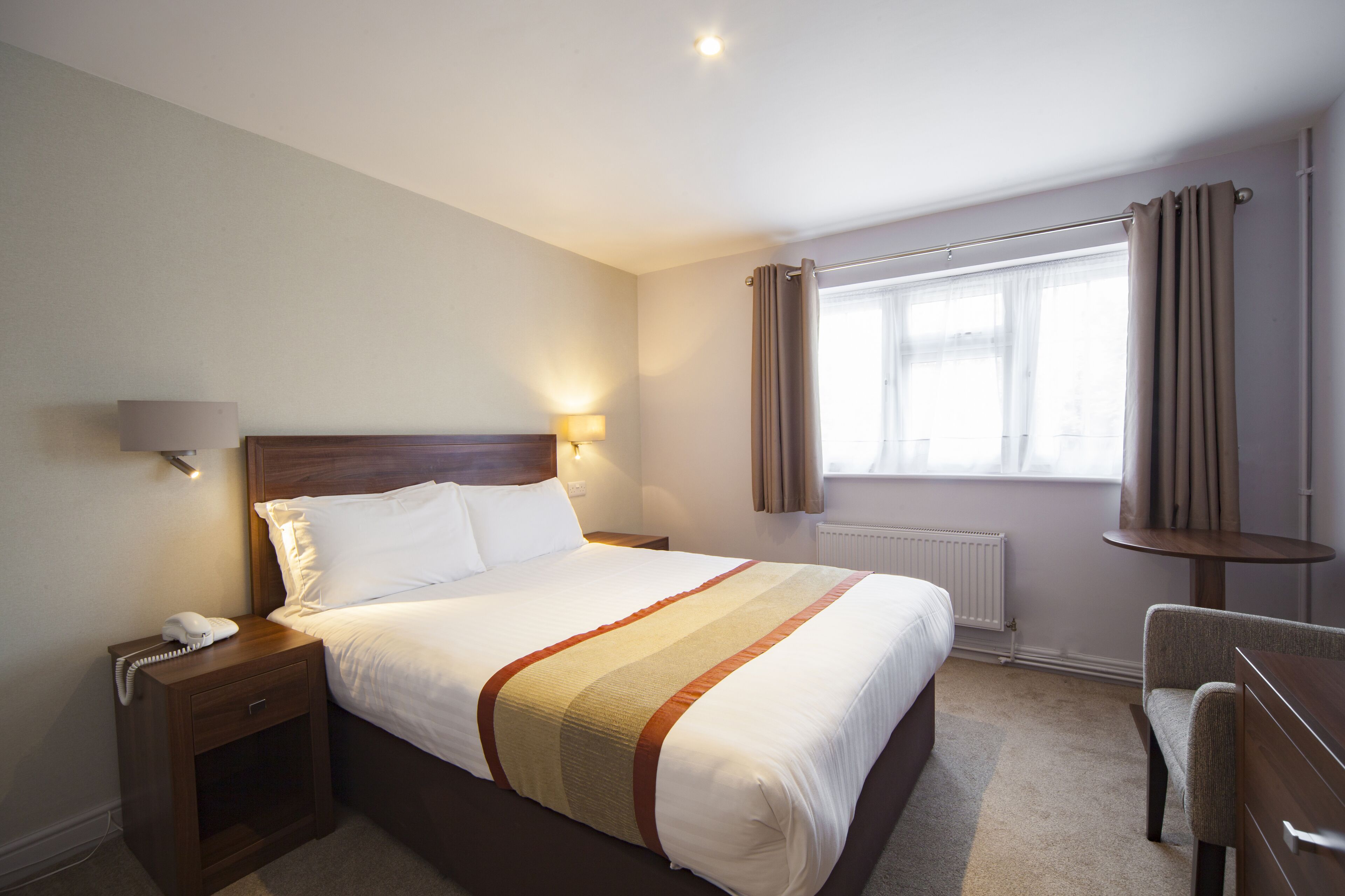A hotel room with a large bed, bedside table with a lamp, a chair, and a window with curtains.