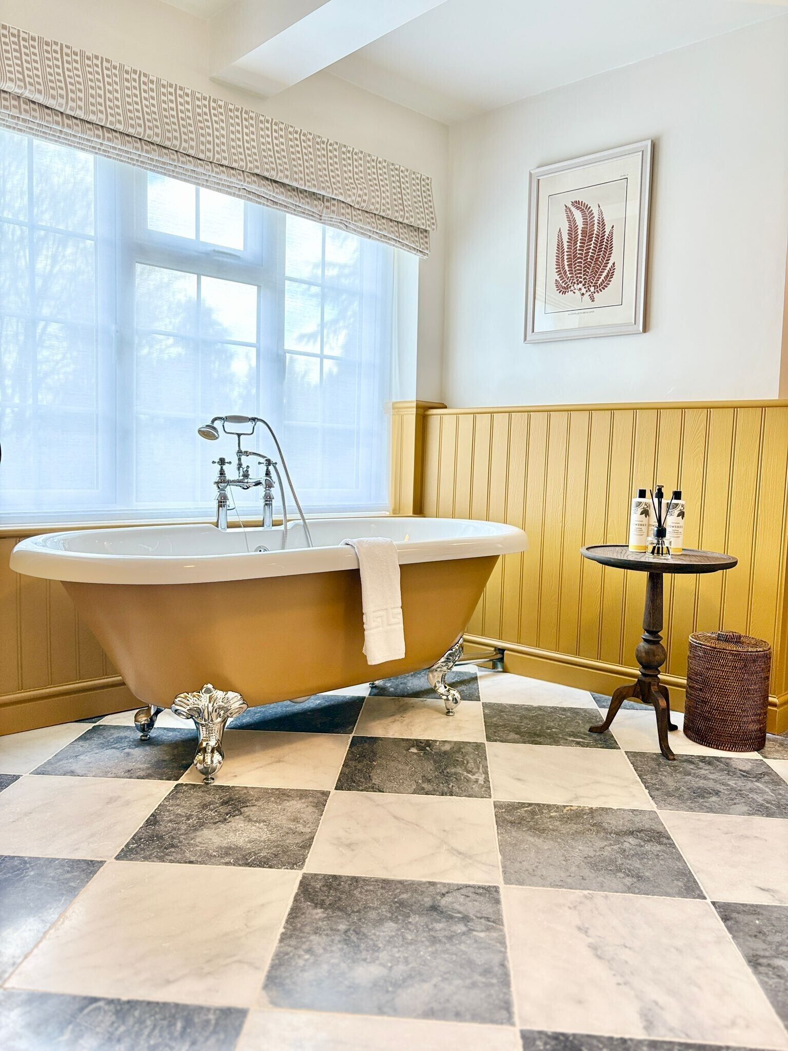A bathroom with a freestanding bathtub, a small round table with toiletries, a framed artwork on the wall, and a window with a patterned valance.
