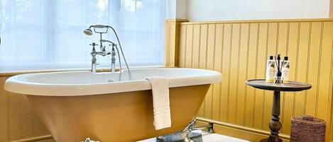 Une salle de bain avec une baignoire sur pieds, une petite table ronde avec des articles de toilette, une œuvre d’art encadrée au mur et une fenêtre agrémentée d’une lambrequin à motifs.