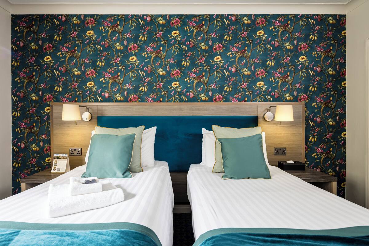 A hotel room with two beds, a floral wallpaper, and bedside lamps.