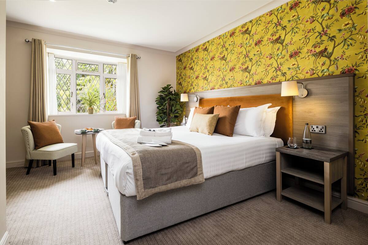 Quarto de hotel com cama grande, mesa de cabeceira, cadeira, mesinha e janela com cortinas.