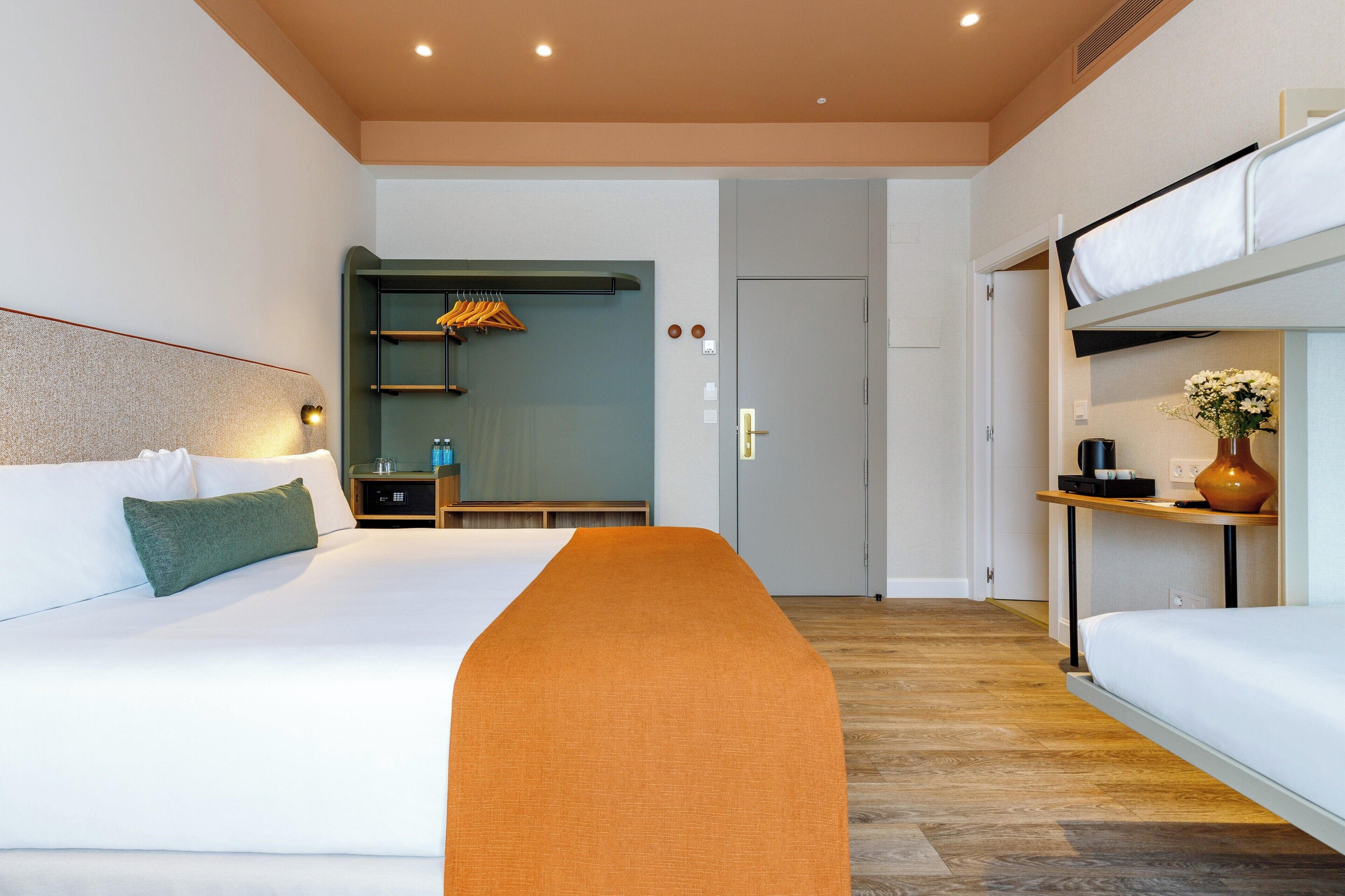 Una habitación de hotel moderna con una cama grande, un banco, una mesita con un jarrón y una cama litera.