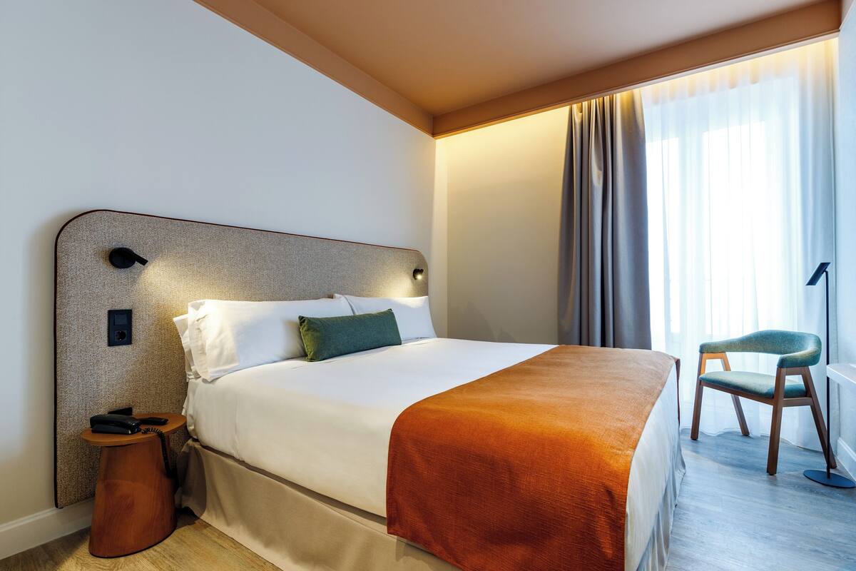 Ein Hotelzimmer mit einem Bett, einem Nachttisch mit Telefon, einem Sessel und einem Fenster mit Vorhängen.