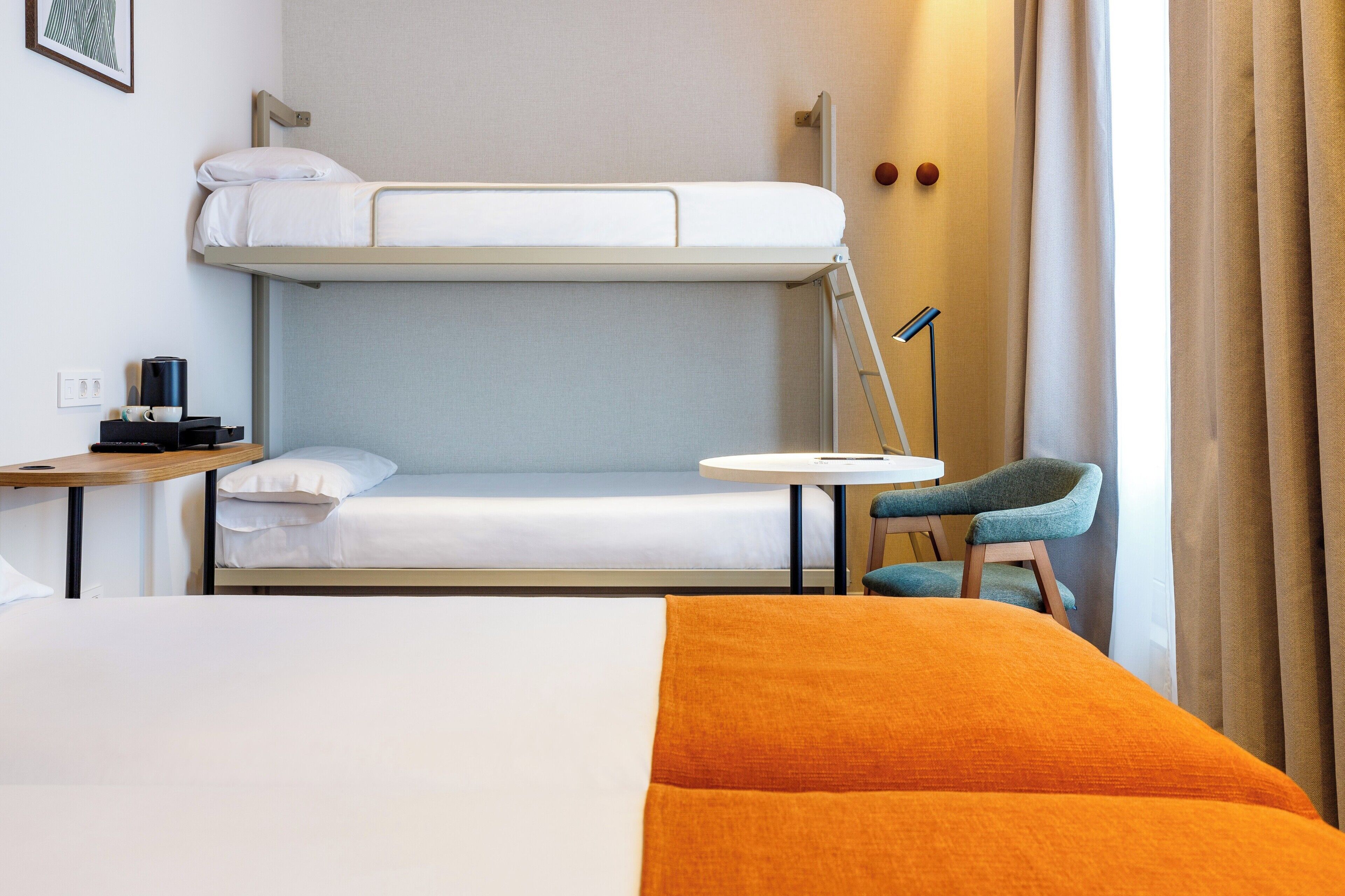 Habitació d'hotel amb un llit superposat, un llit individual, una taula petita amb una làmpada, una cadira i una finestra amb cortines.