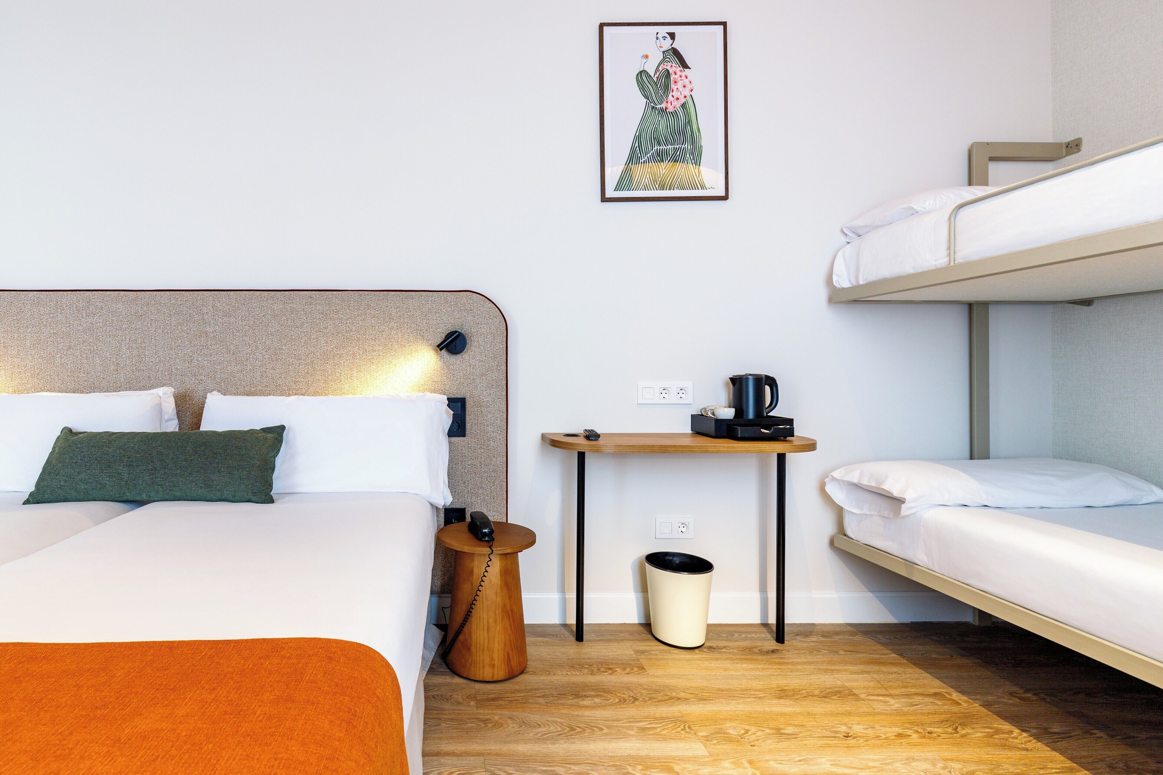 A hotel room with a single bed, a bunk bed, a small wooden table with a coffee maker, a trash bin, and a framed picture on the wall.