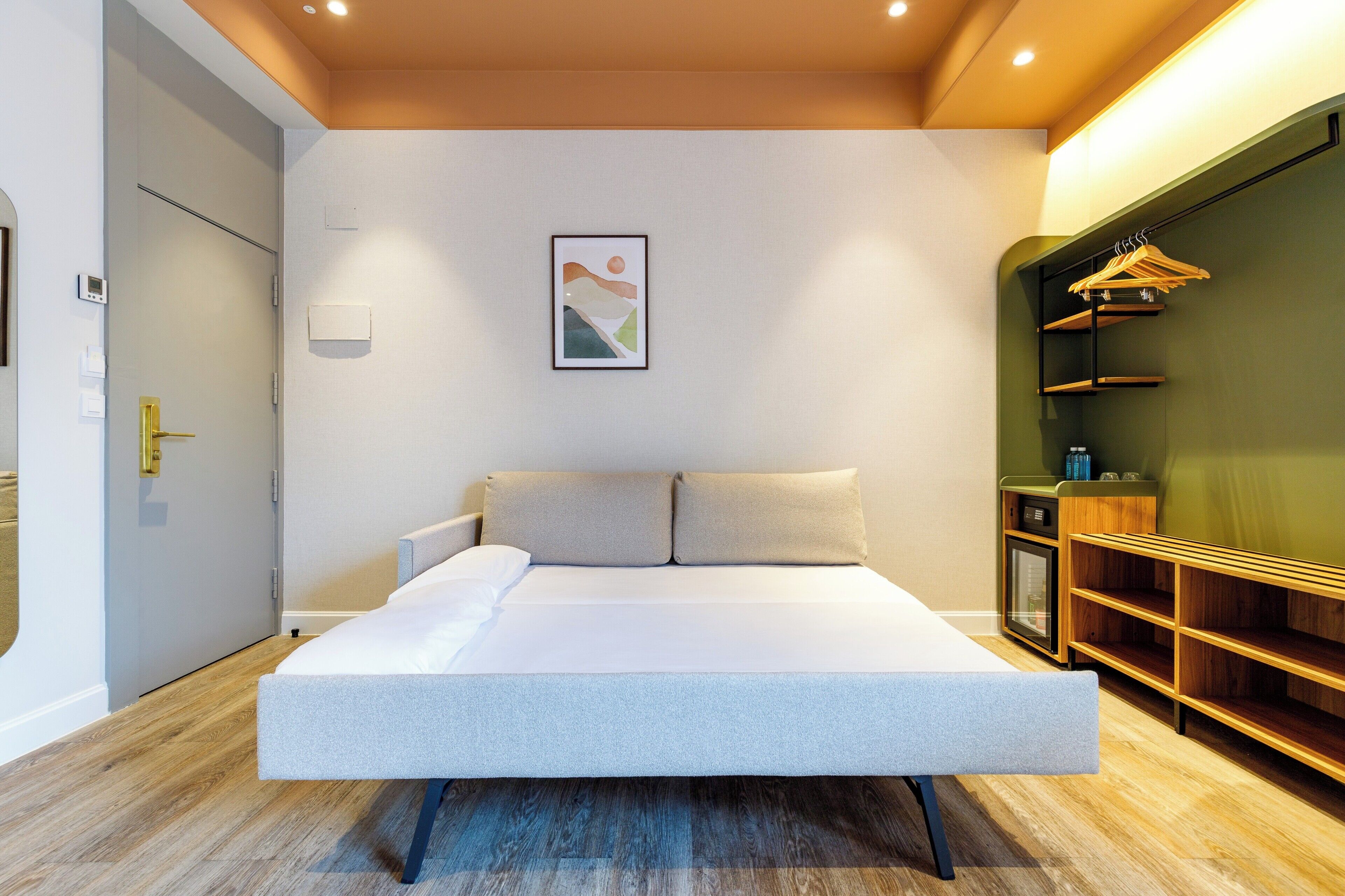 A modern bedroom with a large bed, a bedside table, a wall-mounted shelf with clothes, and a small cabinet.