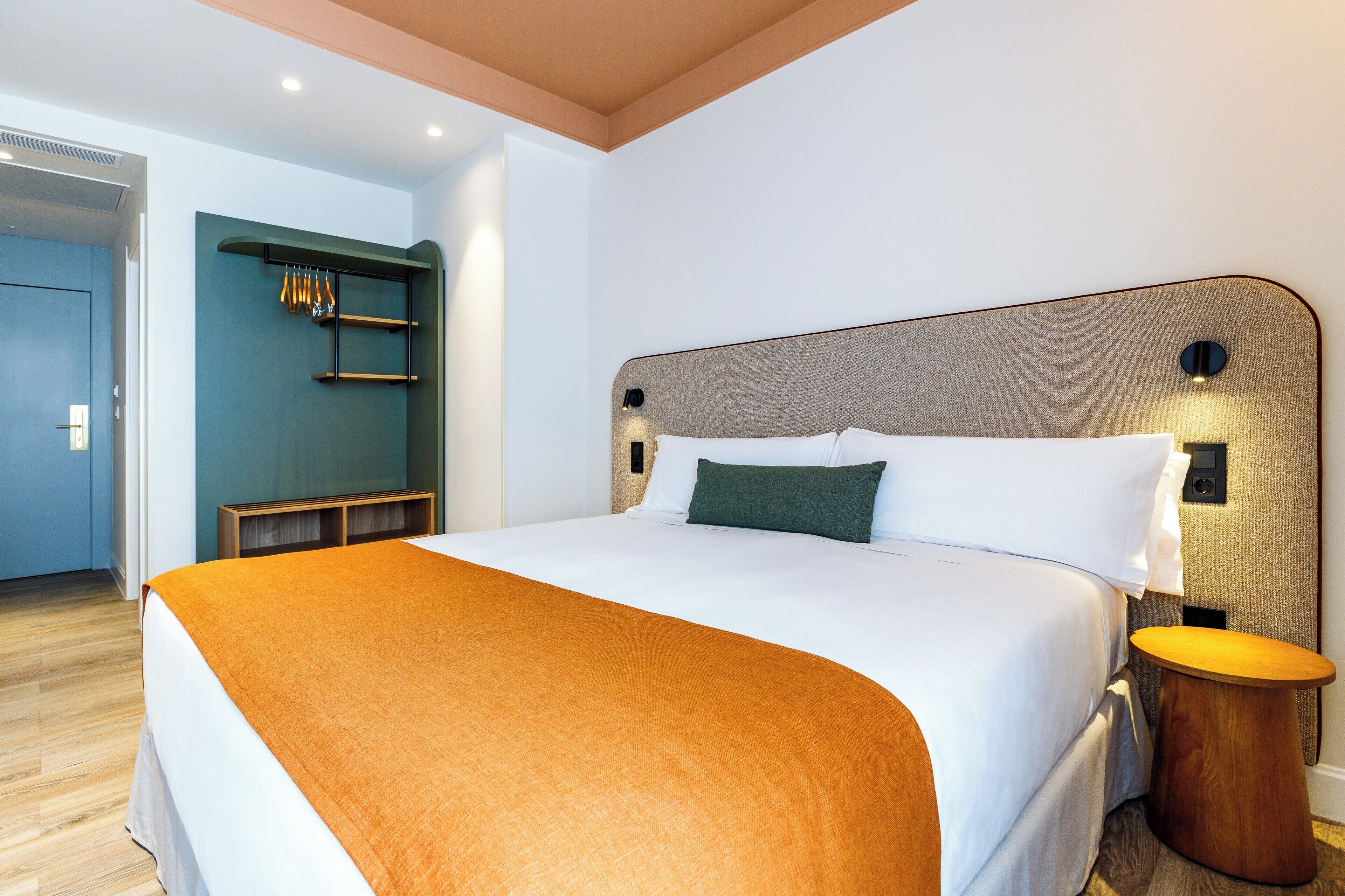 Une chambre d’hôtel avec un grand lit, une table de chevet en bois, une étagère murale avec des objets suspendus et une porte donnant sur une autre pièce.