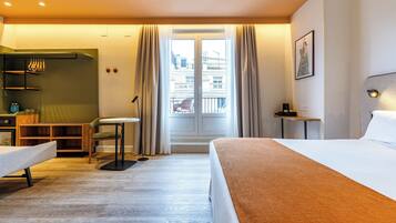A modern hotel room with a large bed, a desk, a chair, and a view of the city through a large window.