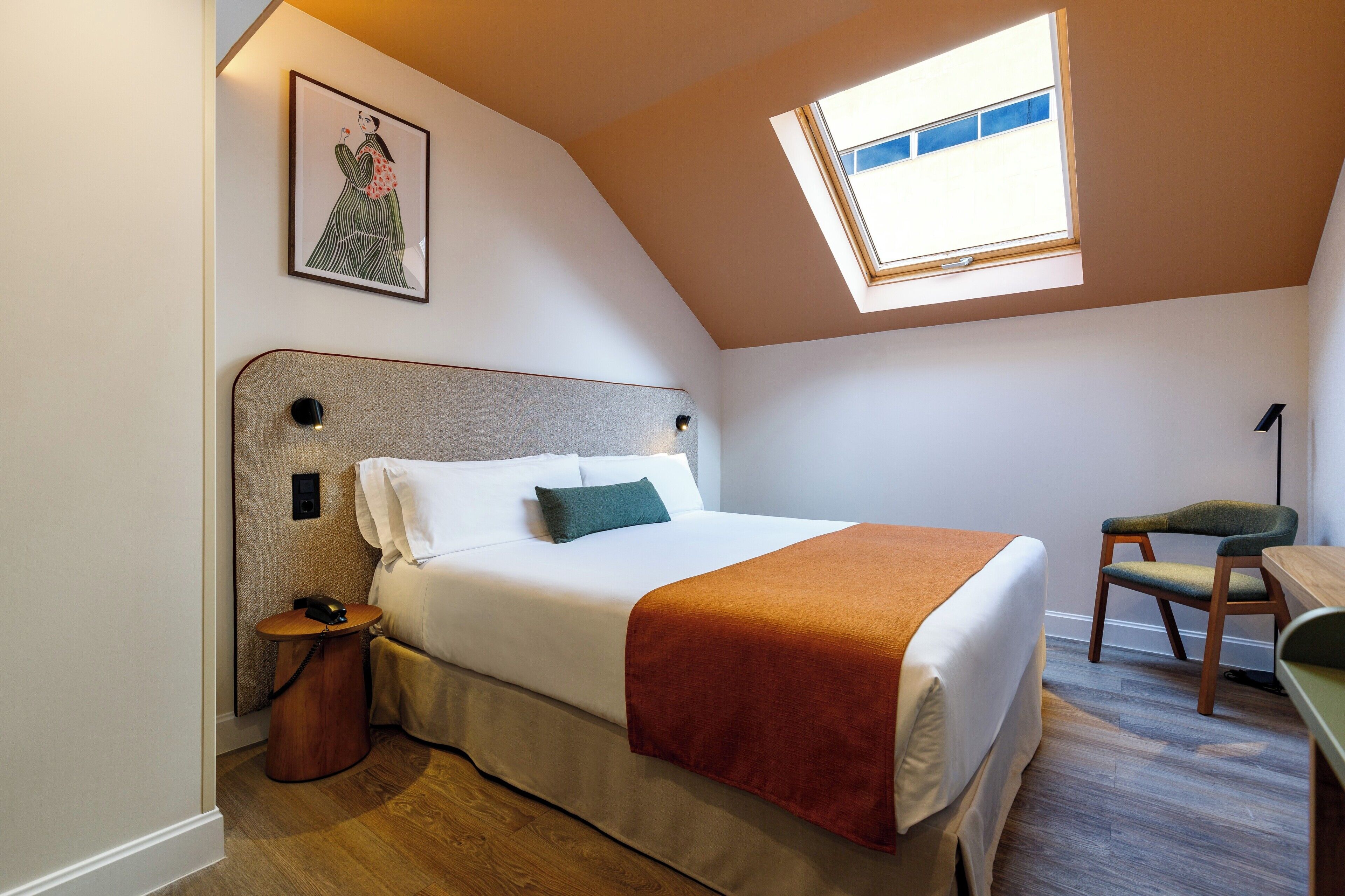 Ein Schlafzimmer mit Bett, Schreibtisch, Stuhl, kleinem Tisch, Wandtelefon, gerahmtem Bild und Oberlicht.