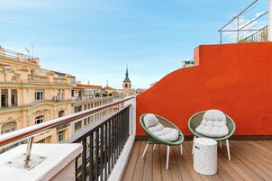 A balcony with two wicker chairs and a small table, overlooking a cityscape with historic buildings and a church spire.