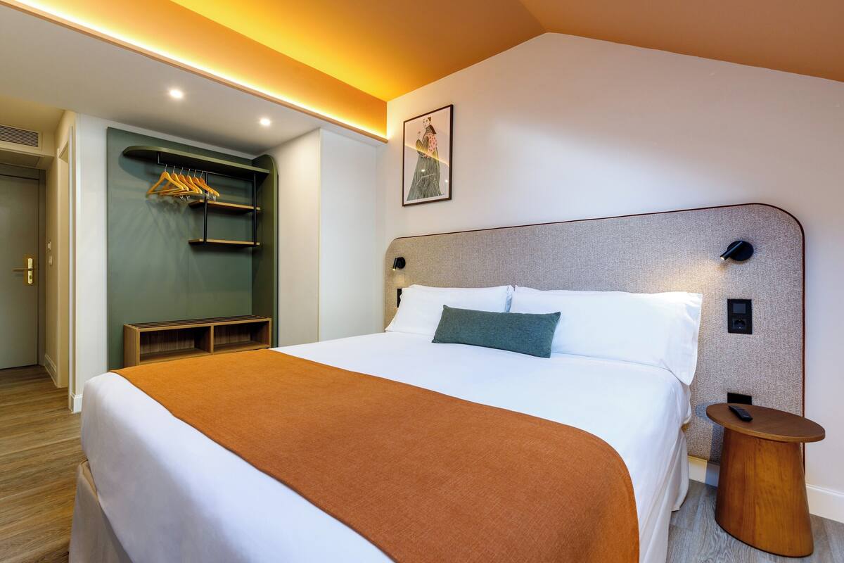 Ein ordentlich gemachtes Bett mit brauner Decke, ein Nachttisch mit Fernbedienung und ein an der Wand hängendes Bild.