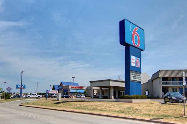 A motel with a prominent '6' sign, a visible entrance, and a 'NOW OPEN' sign. - Motel 6 Mount Pleasant, TX (Mount Pleasant)