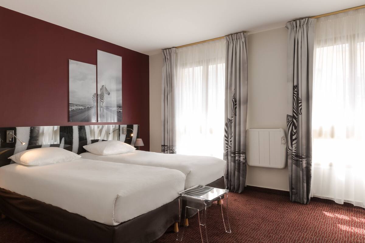 A hotel room with two beds, a small table, a radiator, and curtains.