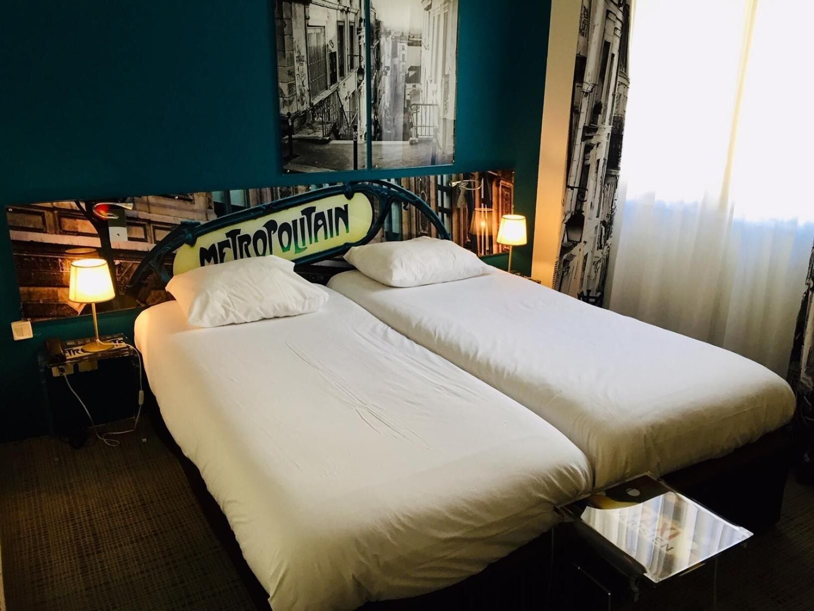 A hotel room with two single beds, a nightstand with a lamp, a small table, and a wall with a cityscape poster.