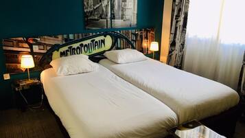 A hotel room with two single beds, a nightstand with a lamp, a small table, and a wall with a cityscape poster.