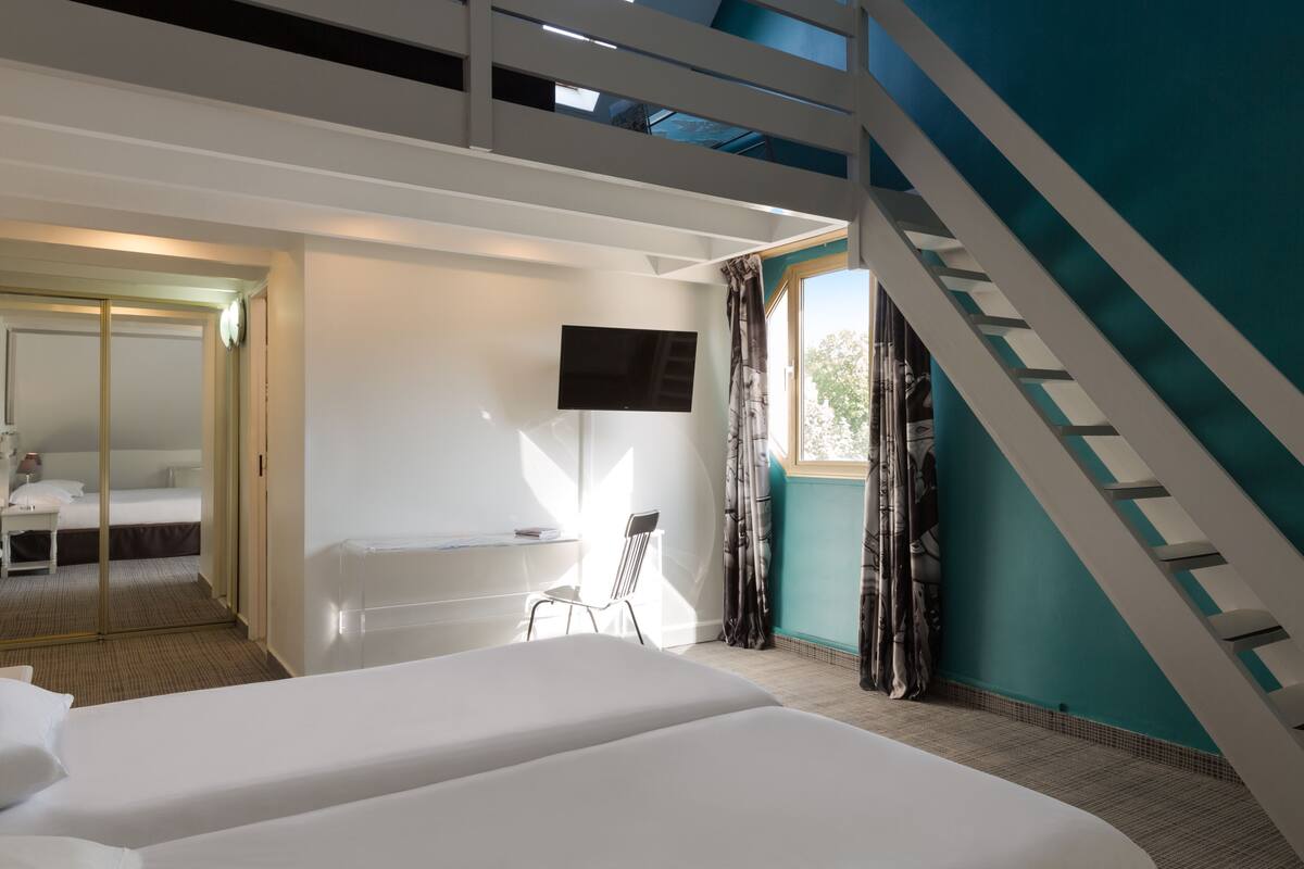 A hotel room with two beds, a TV mounted on the wall, a chair, and a view of the outdoors through a window.