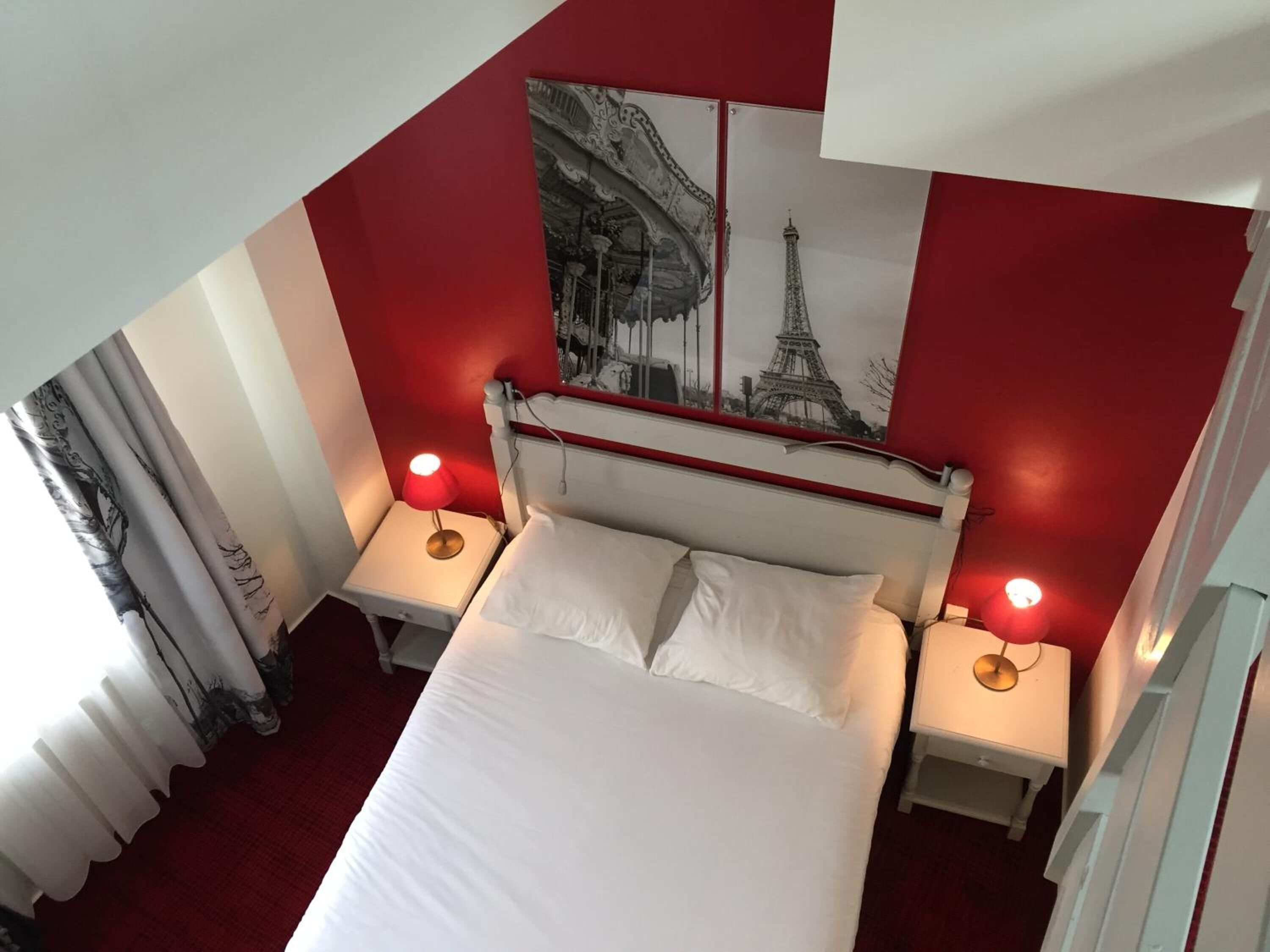 A hotel room with a large bed, two bedside tables with lamps, a red accent wall, and a poster of the Eiffel Tower.