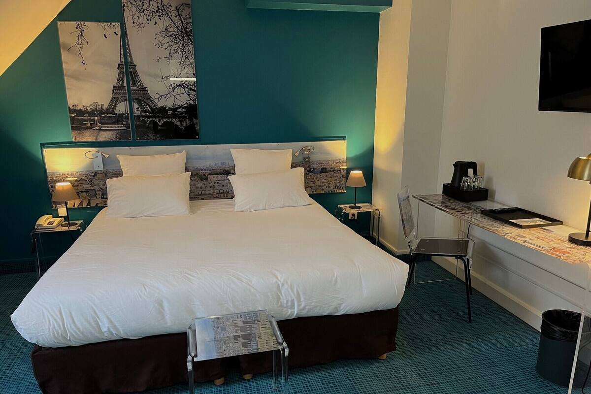 A hotel room with a large bed, a desk, a chair, a trash bin, and a wall-mounted TV.