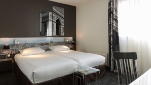 Une chambre d’hôtel avec un grand lit, une table de chevet avec une lampe, une chaise et un tableau représentant un paysage urbain accroché au mur.