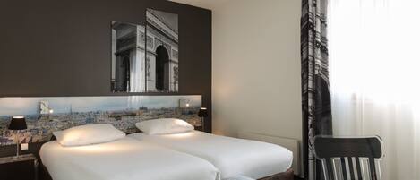 Habitación de hotel con una cama grande, una mesita de noche con lámpara, una silla y un cuadro de un paisaje urbano en la pared.