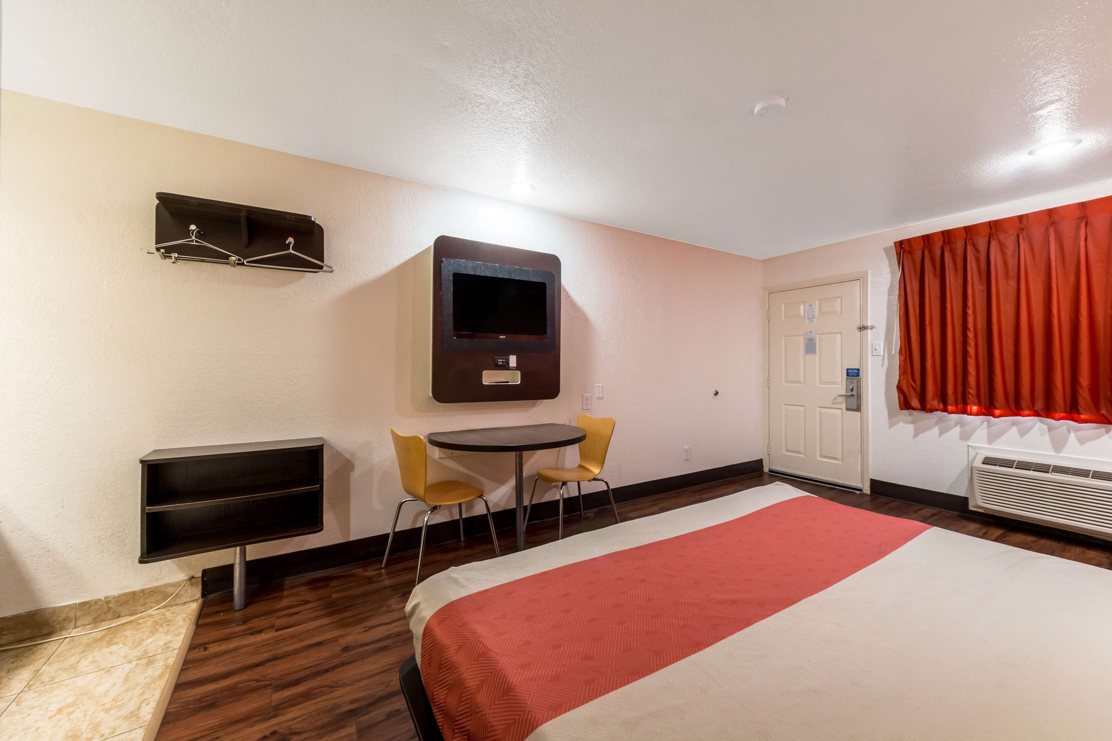 Una habitación de hotel con cama, televisor montado en la pared, escritorio con dos sillas y perchero.
